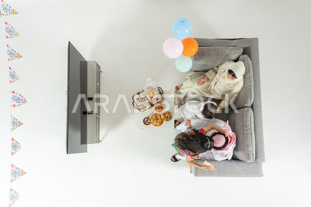 Sharing the Eid atmosphere with the family, a girl kissing her father’s head, a vertical overhead portrait of a Saudi Gulf Arab family wearing the traditional dress sitting on the sofa in the living room, wearing new Eid clothes, presenting Eid gifts to the children, showing a blank black screen on the TV, white background