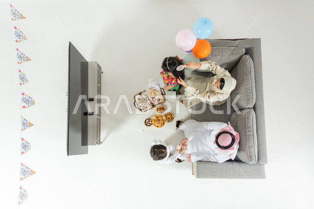 تقديم العيديات للأبناء , بورتريه علوي عامودي لعائلة عربية خليجية سعودية يرتدون الثوب التقليدي يجلسون على الأريكة في غرفة المعيشة , أجواء العيد في السعودية, عرض شاشة سوداء فارغة عبر التلفاز , خلفية بيضاء