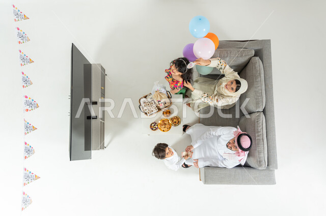 أجواء العيد في السعودية , بورتريه علوي عامودي لعائلة عربية خليجية سعودية يرتدون الثوب التقليدي يجلسون على الأريكة في غرفة المعيشة , تقديم العيديات للأبناء , النظر إلى الكاميرا بإيماءات الفرح و السرور , عرض شاشة سوداء فارغة عبر التلفاز , خلفية بيضاء