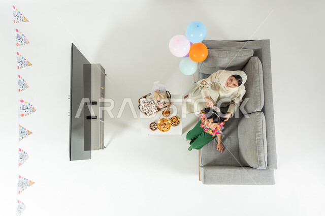 Eid celebration preparations, vertical overhead portrait of a Saudi Arabian Gulf mother wearing a traditional dress, sitting on the sofa in the living room, combing her daughter’s hair, looking at the camera with gestures of joy and happiness, showing a blank black screen on the TV, white background