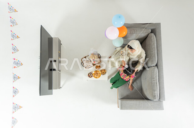 تحضيرات الاحتفال بالمناسبات و الأعياد , بورتريه علوي عامودي لأم عربية خليجية سعودية ترتدي الثوب التقليدي تجلس على الأريكة في غرفة المعيشة تقوم بتمشيط شعر ابنتها , الفرحة بقدوم العيد , عرض شاشة سوداء فارغة عبر التلفاز , خلفية بيضاء