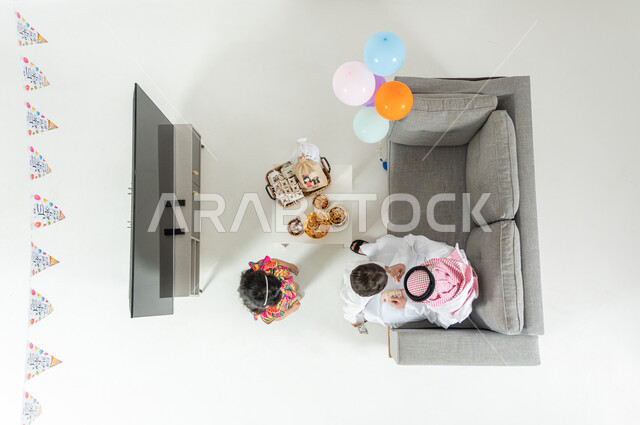 التهنئة بقدوم العيد , بورتريه علوي عامودي لأب عربي خليجي سعودي يرتدي الثوب التقليدي و الشماغ يجلس على الأريكة في غرفة المعيشة يقوم بتقديم العيدية لأبنائه  , أجواء عائلية سعيدة , عرض شاشة سوداء فارغة عبر التلفاز , خلفية بيضاء