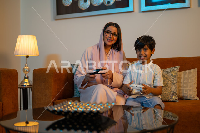 Gestures of happiness with fun recreational activities, entertainment and amusement with video games, a Saudi Arabian Gulf boy holding a controller in his hand and playing PlayStation with his mother, the importance of children sharing leisure time, developing children’s motor and mental abilities