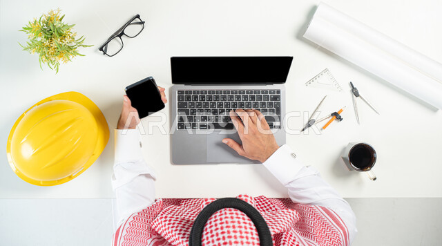 البيع والشراء عبر الانترنت، استخدام المواقع الالكترونية للبحث عن مستلزمات البناء، بورتريه مقرب ليدي رجل عربي خليجي سعودي يستخدم جهاز الهاتف المحمول واللابتوب وبجانبه فنجان من القهوة وأوراق وأدوات هندسية مختلفة، عرض شاشة سوداء فارغة، خلفية بيضاء