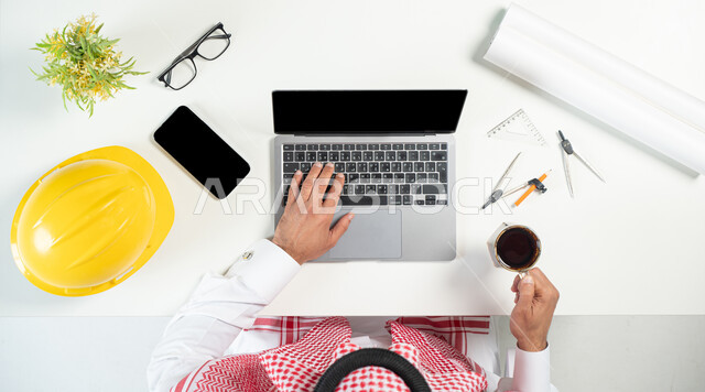 استخدام المواقع الالكترونية للبحث عن مستلزمات البناء، بورتريه مقرب ليدي رجل عربي خليجي سعودي يستخدم جهاز الحاسوب المحمول وبجانبه هاتف محمول وأدوات هندسية، البيع والشراء عبر الانترنت، الاستمتاع بالعمل شرب فنجان من القهوة، عرض شاشة سوداء فارغة، خلفية بيضاء