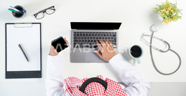 البحث عن استشارات طبية أونلاين من خلال استخدام المواقع الالكترونية، بورتريه مقرب ليدي رجل عربي خليجي سعودي يستخدم جهاز الحاسوب المحمول والجوال وبجانبه فنجان من القهوة وسماعات الأذن للفحص الطبي وأوراق كتابة الوصفات الأدوية اللزمة، عرض شاشة سوداء فارغة، خلفية بيضاء