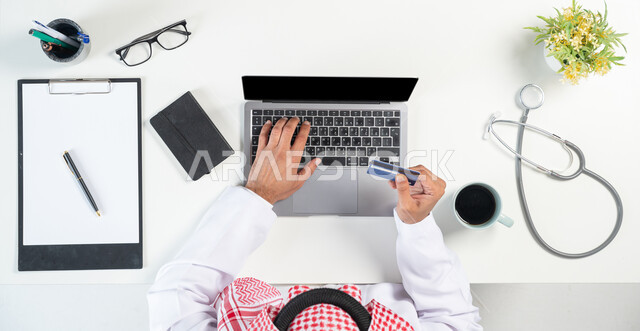 البحث عن استشارة طبية أونلاين من خلال استخدام المواقع الالكترونية، بورتريه مقرب ليدي رجل عربي خليجي سعودي يستخدم جهاز الحاسوب المحمول ويقوم بالدفع من خلال بطاقة الصراف البنكية وبجانبه فنجان من القهوة وسماعات الفحص الطبية، عرض شاشة سوداء فارغة، خلفية بيضاء