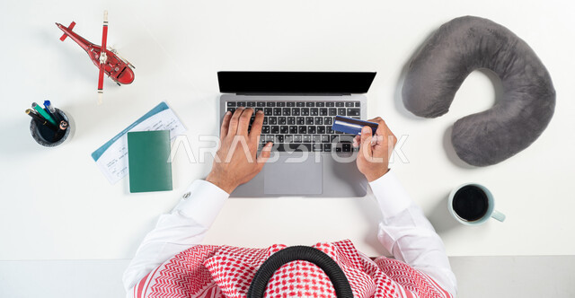 قطع تذاكر الطيران أونلاين، استخدام المواقع الالكترونية للبحث عن رحلات جوية، بورتريه مقرب ليدي رجل عربي خليجي سعودي يستخدم جهاز الحاسوب المحمول للدفع عبر بطاقة الصراف البنكية وبجانبه جواز وتذكرة السفر وفنجان من القهوة ومجسمات مصغرة للطائرات، عرض شاشة سوداء فارغة، خلفية بيضاء