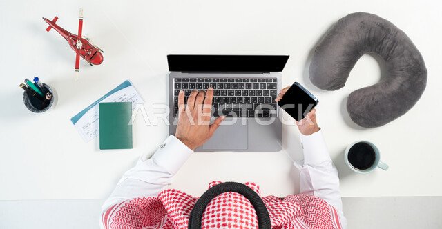 قطع تذاكر الطيران أونلاين، استخدام المواقع الالكترونية للبحث عن رحلات جوية، بورتريه مقرب ليدي رجل عربي خليجي سعودي يستخدم جهاز الحاسوب المحمول والجوال  وبجانبه جواز وتذكرة السفر وفنجان من القهوة ومجسمات مصغرة للطائرات، البيع والشراء عبر الانترنت، عرض شاشة سوداء فارغة، خلفية بيضاء