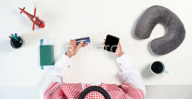 قطع تذاكر الطيران أونلاين، استخدام المواقع الالكترونية للبحث عن رحلات جوية، بورتريه مقرب ليدي رجل عربي خليجي سعودي يستخدم جهاز الهاتف المحمول للدفع عن طريق بطاقة الصراف البنكية وبجانبه فنجان من القهوة ومجسمات مصغرة للطائرات، عرض شاشة سوداء فارغة، خلفية بيضاء