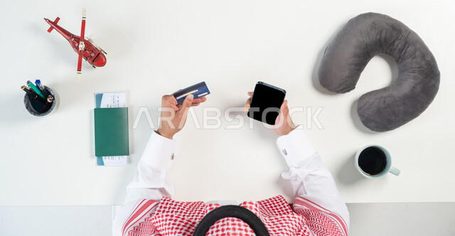 قطع تذاكر الطيران أونلاين، استخدام المواقع الالكترونية للبحث عن رحلات جوية، بورتريه مقرب ليدي رجل عربي خليجي سعودي يستخدم جهاز الهاتف المحمول للدفع عن طريق بطاقة الصراف البنكية وبجانبه فنجان من القهوة ومجسمات مصغرة للطائرات، عرض شاشة سوداء فارغة، خلفية بيضاء