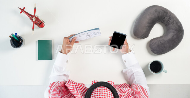 البيع والشراء عبر الانترنت، قطع تذاكر الطيران أونلاين، استخدام المواقع الالكترونية للبحث عن رحلات جوية، بورتريه مقرب ليدي رجل عربي خليجي سعودي يستخدم جهاز الهاتف المحمول ويحمل بيده تذكرة السفر وبجانبه فنجان من القهوة ومجسمات مصغرة للطائرات، عرض شاشة سوداء فارغة، خلفية بيضاء