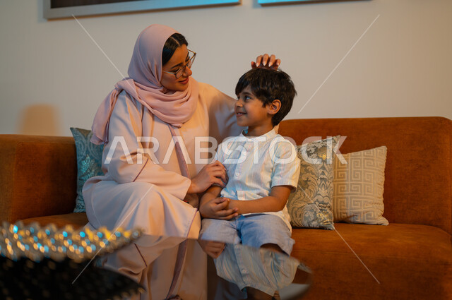 أهمية مشاركة الأبناء أوقات الفراغ، إيماءات السعادة والسرور بقضاء أوقات عائلية ممتعة يوم العطلة، أم عربية خليجية سعودية محجبة ترتدي عباءة عصرية تجلس على الأريكة في غرفة المعيشة برفقة ابنها