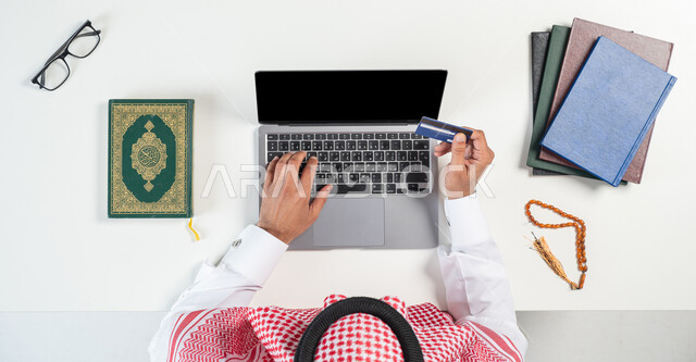 دمج التكنولوجيا بالحياة اليومية، بورتريه ليدي رجل عربي خليجي سعودي يقوم باستخدام الحاسوب المحمول للدفع من خلال بطاقة المصرف البنكية وبجانبه القرآن الكريم ومسبحة صفراء، شراء الكتب الدينية والمصاحف الشريفة من خلال المواقع الالكترونية، عرض شاشة سوداء فارغة، خلفية بيضاء