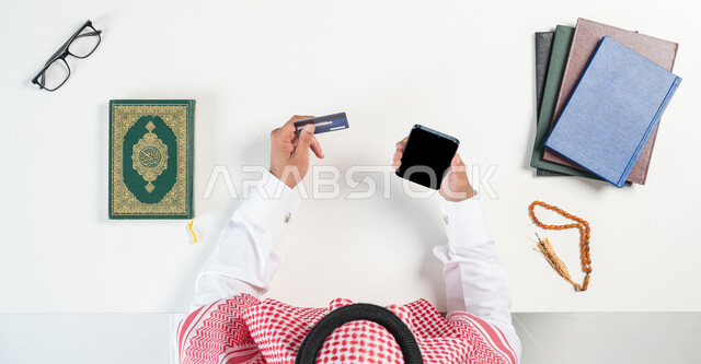 دمج التكنولوجيا بالحياة اليومية، بورتريه ليدي رجل عربي خليجي سعودي يقوم باستخدام الهاتف المحمول للدفع من خلال بطاقة المصرف البنكية وبجانبه القرآن الكريم ومسبحة صفراء، شراء الكتب الدينية والمصاحف الشريفة من خلال المواقع الالكترونية، عرض شاشة سوداء فارغة، خلفية بيضاء