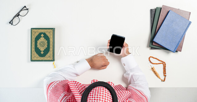 البيع والشراء عبر الانترنت، دمج التكنولوجيا بالحياة اليومية، بورتريه ليدي رجل عربي خليجي سعودي يقوم باستخدام الهاتف المحمول وبجانبه القرآن الكريم ومسبحة صفراء، شراء الكتب الدينية والمصاحف الشريفة من خلال المواقع الالكترونية، عرض شاشة سوداء فارغة، خلفية بيضاء