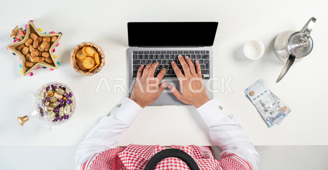 الاحتفال بالمناسبات والأعياد، تبادل التهنئة وارسال العيادي من خلال الانترنت، بيع وشراء ضيافة العيد أونلاين، بورتريه مقرب ليدي رجل عربي خليجي سعودي يقوم باستخدام الحاسوب المحمول وبجانبه طبق من التمر وأنواع من الحلويات،عرض شاشة سوداء فارغة، خلفية بيضاء