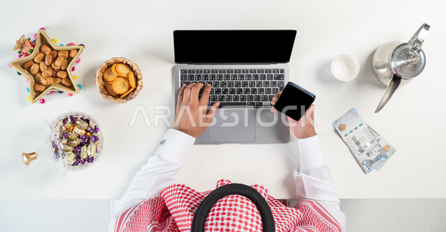 الاحتفال بالمناسبات والأعياد، تبادل التهنئة وارسال العيادي من خلال الانترنت، بيع وشراء ضيافة العيد أونلاين، بورتريه مقرب ليدي رجل عربي خليجي سعودي يقوم باستخدام اللابتوب وبيده الهاتف المحمول وبجانبه طبق من التمر وأنواع من الحلويات،عرض شاشة سوداء فارغة، خلفية بيضاء