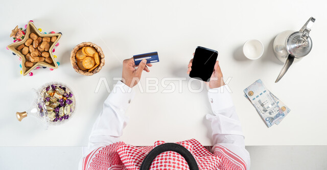 Exchanging congratulations and sending Eid holidays online, buying and selling Eid hospitality online, a close-up portrait of a Saudi Gulf Arab man using a mobile phone to pay via a bank ATM card, next to him is a plate of dates and various types of sweets, displaying a blank black screen, white background