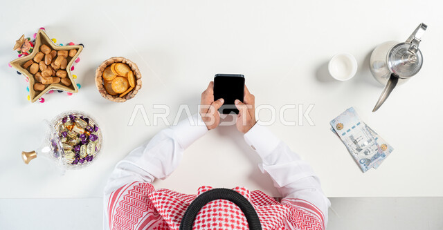 بيع وشراء ضيافة العيد أونلاين، تبادل التهنئة بمناسبة العيد من خلال استخدام المواقع الالكترونية، بورتريه مقرب ليدي رجل عربي خليجي سعودي يقوم بحمل واستخدام الجوال وبجانبه طبق من التمر وأنواع من الحلويات دلة من القهوة، ارسال العيديات من خلال الانترنت، خلفية بيضاء