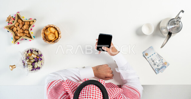 بيع وشراء ضيافة العيد أونلاين، تبادل التهنئة بمناسبة العيد من خلال استخدام المواقع الالكترونية، بورتريه مقرب ليدي رجل عربي خليجي سعودي يقوم باستخدام الجوال وبجانبه طبق من التمر وأنواع من الحلويات دلة من القهوة، ارسال العيديات من خلال الانترنت، خلفية بيضاء