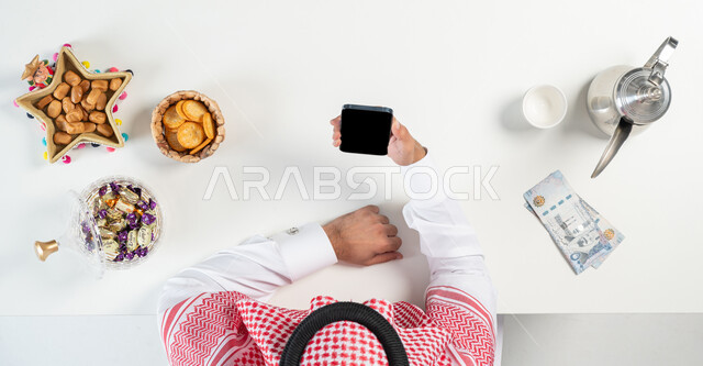 بيع وشراء ضيافة العيد أونلاين، تبادل التهنئة بمناسبة العيد من خلال استخدام المواقع الالكترونية، بورتريه مقرب ليدي رجل عربي خليجي سعودي يقوم باستخدام الجوال وبجانبه طبق من التمر وأنواع من الحلويات دلة من القهوة، ارسال العيديات من خلال الانترنت، خلفية بيضاء