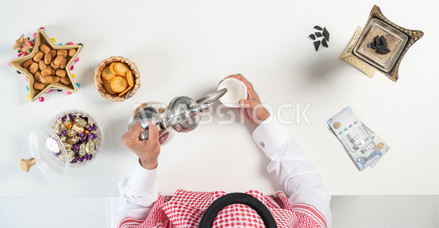 تبادل التهنئة وتقديم العيادي بمناسبة العيد، تقديم تشكيلة متنوعة من المأكولات الغنية بالسكريات والسعرات الحرارية، بورتريه مقرب ليدي رجل عربي خليجي سعودي يقوم بصب فنجان من القهوة المرة وبجانبه طبق من التمر وأنواع من الحلويات، خلفية بيضاء