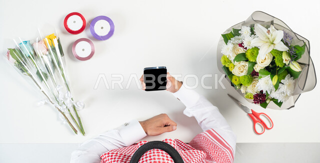 صناعة وبيع وشراء بوكيه الورد عبر الانترنت، بورتريه ليدي رجل عربي خليجي سعودي يقوم باستخدام الهاتف المحمول للحصول على أدوات الأزهار عن طريق الأونلاين، استخدام التكنولوجيا في التعلم والتدريب من خلال المواقع الإلكترونية، عرض شاشة سوداء فارغة، خلفية بيضاء