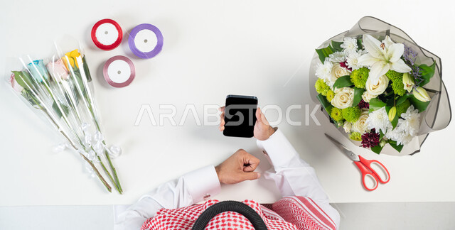 صناعة وبيع وشراء بوكيه الورد عبر الانترنت، بورتريه ليدي رجل عربي خليجي سعودي يقوم باستخدام الهاتف المحمول للحصول على أدوات الأزهار عن طريق الأونلاين، استخدام التكنولوجيا في التعلم والتدريب من خلال المواقع الإلكترونية، عرض شاشة سوداء فارغة، خلفية بيضاء