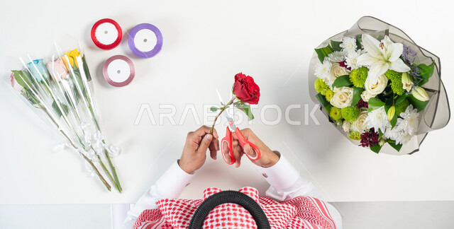 صنع هدايا للتعبير عن الحب، تعلم كيفية تحضير وتجهيز بوكيه الزهور، بورتريه ليدي رجل عربي خليجي سعودي يقوم باستخدام مقص تقليم الورود وبجانبه مجموعة من الأزهار الملونة، خلفية بيضاء