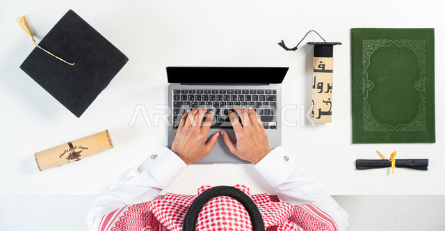 Using remote shopping applications, taking advantage of modern technology to order graduation supplies online, portrait of a Saudi Gulf Arab man using a laptop with documents and a graduation cap next to him, buying and selling online, displaying a blank black screen, white background