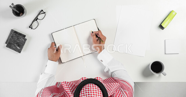 تدوين الملاحظات والأفكار وانجاز مهام العمل، بورتريه ليدي رجل عربي خليجي سعودي يقوم بالكتابة على دفتر وبجانبه فنجان من القهوة ونظارات طبية وأدوات القرطاسية على الطاولة، خلفية بيضاء