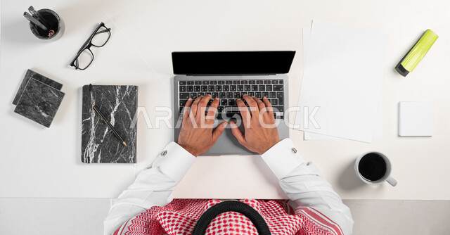 البيع والشراء باستخدام الأجهزة الالكترونية أونلاين، الاستمتاع بشرب فنجان القهوة أثناء العمل، بورتريه ليدي رجل عربي خليجي سعودي يقوم باستعمال الحاسوب المحمول وبجانبه مجموعة من الأدوات المكتبية والقرطاسية على الطاولة، عرض شاشة سوداء فارغة، خلفية بيضاء