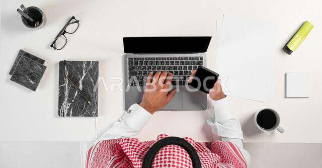 الاستمتاع بشرب فنجان القهوة أثناء العمل، بورتريه ليدي رجل عربي خليجي سعودي يقوم باستعمال الحاسوب المحمول والجوال وبجانبه مجموعة من الأدوات المكتبية والقرطاسية على الطاولة، البيع والشراء باستخدام الأجهزة الالكترونية أونلاين، عرض شاشة سوداء فارغة، خلفية بيضاء