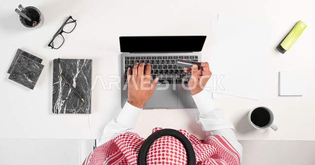 Enjoying drinking a cup of coffee while working, portrait of a Saudi Gulf Arab man using a laptop to pay via a bank debit card, next to him a group of office supplies and stationery on the table, buying and selling using electronic devices online, displaying a blank black screen, white background