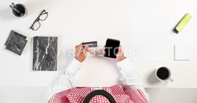 الاستمتاع بشرب فنجان القهوة أثناء العمل، بورتريه ليدي رجل عربي خليجي سعودي يقوم باستعمال الجوال للدفع عبر بطاقة الصراف البنكية، وبجانبه مجموعة من الأدوات المكتبية والقرطاسية على الطاولة، البيع والشراء باستخدام الأجهزة الالكترونية أونلاين، عرض شاشة سوداء فارغة، خلفية بيضاء