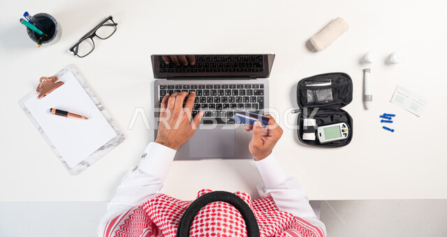 طلب استشارة صحية عبر التطبيقات الالكترونية، الحصول على المستلزمات والأدوات الطبية من خلال الجوال، بورتريه مقرب ليدي رجل عربي خليجي سعودي يستخدم الحاسوب المحمول للدفع عبر بطاقة الصراف البنكية وبجانبه جهاز قياس سكر الدم، البيع والشراء أونلاين، عرض شاشة سوداء فارغة، خلفية بيضاء