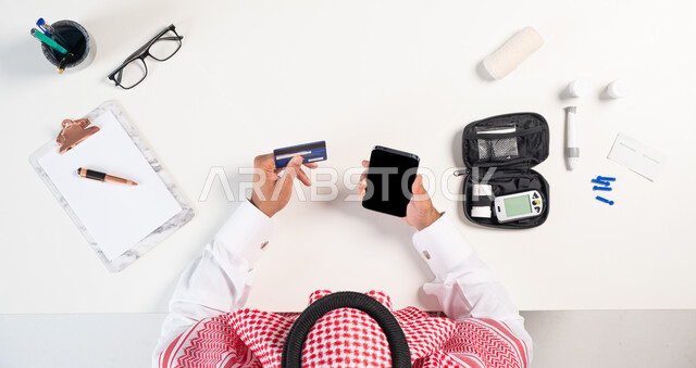 طلب استشارة صحية عبر التطبيقات الالكترونية، الحصول على المستلزمات والأدوات الطبية من خلال الجوال، بورتريه مقرب ليدي رجل عربي خليجي سعودي يستخدم الهاتف المحمول للدفع عبر بطاقة الصراف البنكية وبجانبه جهاز قياس سكر الدم،  البيع والشراء أونلاين، عرض شاشة سوداء فارغة، خلفية بيضاء