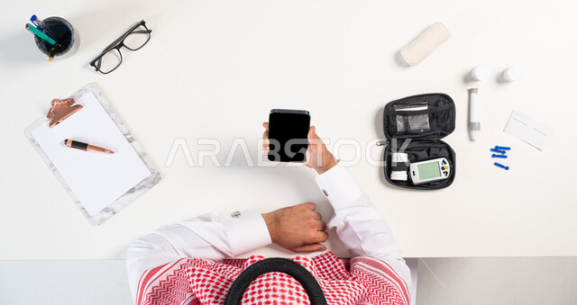 الحصول على المستلزمات والأدوات الطبية من خلال الجوال، بورتريه مقرب ليدي رجل عربي خليجي سعودي يستخدم الهاتف المحمول لطلب استشارة صحية عبر التطبيقات الالكترونية وبجانبه جهاز قياس سكر الدم، بيع وشراء إبر الوخز لتحليل الدم أونلاين، عرض شاشة سوداء فارغة، خلفية بيضاء