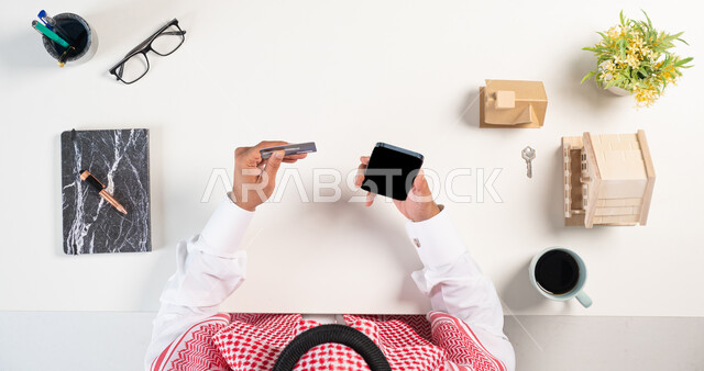 الحصول على المنازل عن طريق الأونلاين، بورتريه علوي عامودي لرجل عربي خليجي سعودي يرتدي الثوب التقليدي والشماغ يستخدم الهاتف المحمول بجانبه مجسم لمنزل وفنجان من القهوة وكتاب، الدفع من خلال بطاقة الصراف البنكية، استخدام التكنولوجيا  في طلب المستلزمات، خلفية بيضاء