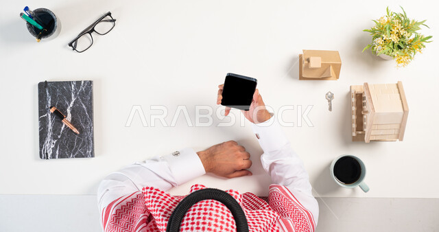 التصفح والتسوق عبر المواقع الالكترونية، بورتريه علوي عامودي لرجل عربي خليجي سعودي يرتدي الثوب التقليدي والشماغ يستخدم الهاتف بجانبه مجسم لمنزل وفنجان من القهوة وكتاب، استخدام التكنولوجيا  في طلب المستلزمات، الحصول على المنازل عن طريق الأونلاين، خلفية بيضاء