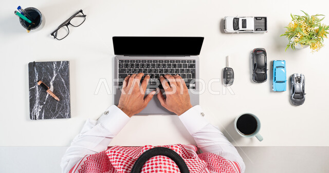 Ordering, buying and selling from the Internet, using technology to order supplies, obtaining modern cars online, a vertical overhead portrait of a Saudi Gulf Arab man wearing a traditional thobe and shemagh, using a laptop computer next to him, a group of cars, a book and a cup of coffee, a white background.