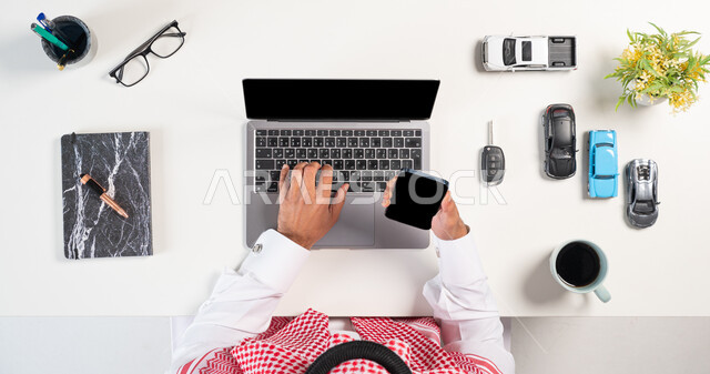 استعمال التقنيات في طلب المستلزمات، الحصول على السيارات الحديثة اونلاين، التسوق عبر المواقع الالكترونية، بورتريه علوي عامودي لرجل عربي خليجي سعودي يرتدي الثوب التقليدي والشماغ يستخدم الهاتف والحاسوب بجانبه مجموعة سيارات وفنجان من القهوة وكتاب، خلفية بيضاء