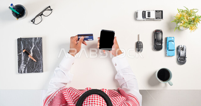 الدفع عن طريق بطاقة المصرف البنكية،  بورتريه علوي عامودي لرجل عربي خليجي سعودي يرتدي الثوب التقليدي والشماغ يستخدم الهاتف بجانبه مجموعة سيارات وفنجان من القهوة وكتاب، استخدام التكنولوجيا  في طلب المستلزمات، الحصول على السيارات الحديثة اونلاين، خلفية بيضاء