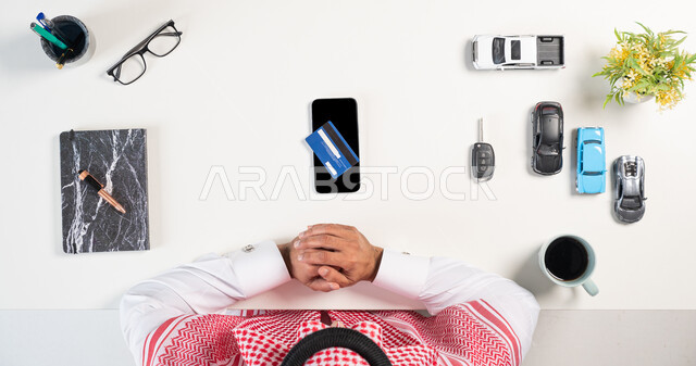 استخدام التكنولوجيا الحديثة، الحصول على السيارات اونلاين، بورتريه علوي عامودي لرجل عربي خليجي سعودي يرتدي الثوب التقليدي والشماغ أمامه الهاتف وبجانبه مجموعة سيارات وفنجان من القهوة وكتاب، الدفع عن طريق بطاقة المصرف البنكية، خلفية بيضاء