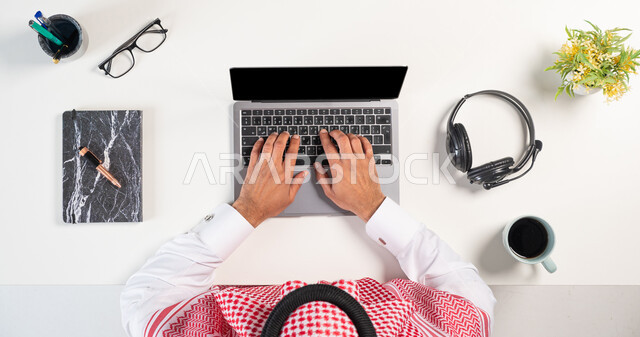 Using modern technology for fun and entertainment, online shopping, using a headphone for technical support, a vertical overhead portrait of a Saudi Gulf Arab man wearing a traditional thobe and shemagh, using a phone and computer with a cup of coffee and a book next to him, white background.