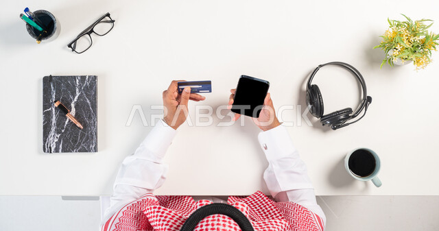 الدفع من خلال بطاقة الصراف البنكية، بورتريه علوي عامودي لرجل عربي خليجي سعودي يرتدي الثوب التقليدي والشماغ يستخدم الهاتف بجانبه فنجان من القهوة وكتاب، مفهوم التسلية والترفيه بالتقنية والتكنولوجيا الحديثة،استخدام الهيدفون للدعم الفني، خلفية بيضاء