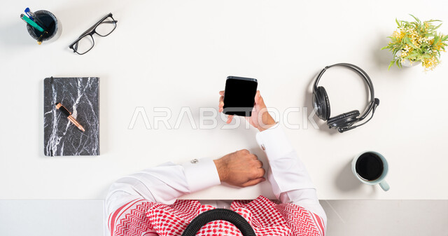 مفهوم  التسلية والترفيه بالتقنية الحديثة، بورتريه علوي عامودي لرجل عربي خليجي سعودي يرتدي الثوب التقليدي والشماغ يستخدم الهاتف بجانبه فنجان من القهوة كتاب، استخدام الهيدفون للدعم الفني، التسوق عبر المواقع الالكترونية، الشراء والبيع عبر الانترنت، خلفية بيضاء