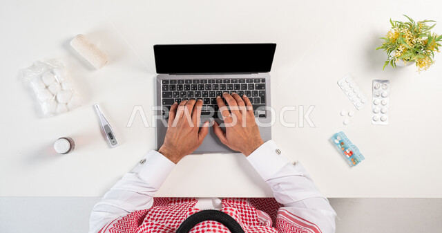 Ordering, buying and selling online, obtaining a variety of medicine, using modern technologies to order pharmaceutical supplies, a vertical overhead portrait of a Saudi Gulf Arab man wearing a traditional thobe and shemagh, using a computer with a group of medicines next to him, white background.