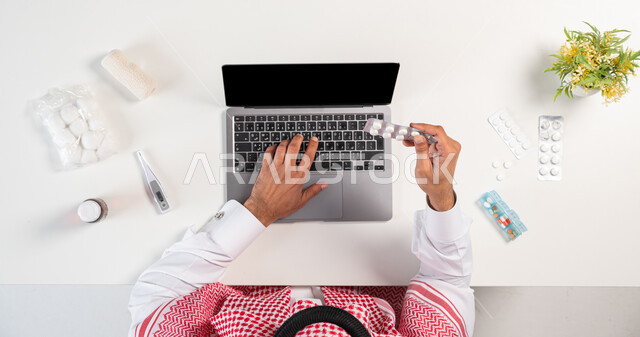 الحصول على مجموعة منوعة من الدواء، استخدام التقنيات الحديثة لطلب المستلزمات الدوائية اونلاين، بورتريه علوي عامودي لرجل عربي خليجي سعودي يرتدي الثوب التقليدي والشماغ يستخدم الحاسوب بجانبه مجموعة من الأدوية، مفهوم التسوق عبر المواقع الالكترونية، خلفية بيضاء